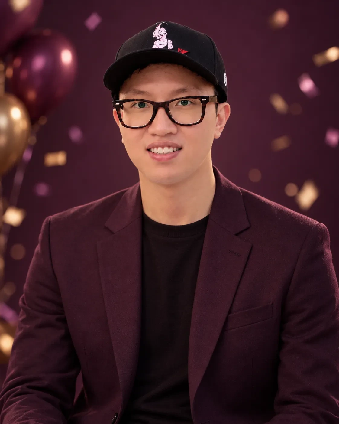 Man in glasses and a cap wearing a burgundy blazer over a black top against a deep plum backdrop with balloons and confetti bokeh