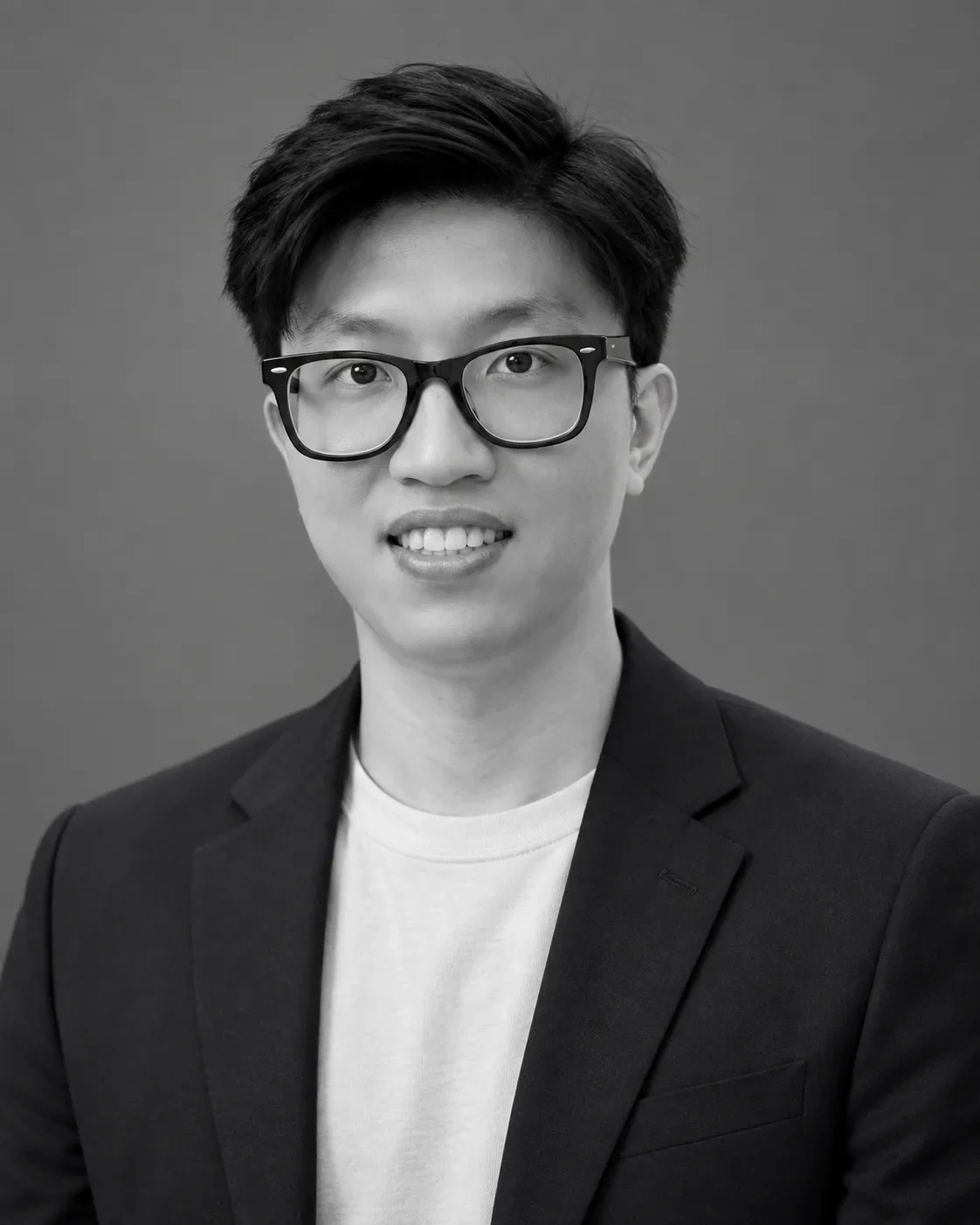 Black-and-white chest-up headshot of a man in glasses wearing a dark blazer over a white T-shirt on a gray backdrop